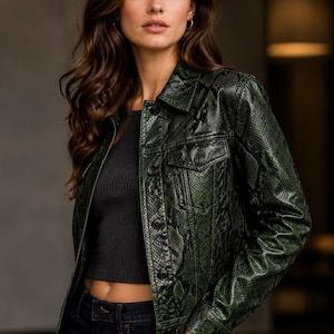 May include: A dark green jacket featuring a textured snakeskin pattern, a classic collar, long sleeves, and a button-down front closure. The garment is worn over a simple black top and dark blue denim jeans, showcasing a structured fit.