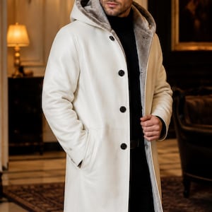 May include: A long, white leather coat featuring a hood, a soft grey fur-like lining, and four large black buttons down the front. The coat has long sleeves and side pockets, worn over a black turtleneck sweater and black trousers.
