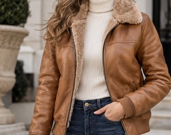 May include: A tan leather bomber jacket featuring a plush beige faux-fur lining and collar, a front zipper closure, and ribbed knit cuffs. The jacket is worn over a cream-colored ribbed turtleneck sweater paired with dark blue denim jeans.