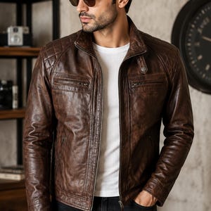 May include: A brown leather jacket featuring a quilted shoulder design, a stand-up collar with a snap button, and two horizontal zippered chest pockets. The jacket is worn over a plain white t-shirt and paired with dark-colored denim jeans.