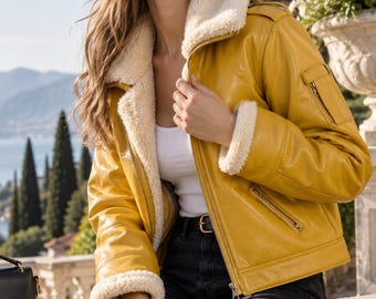 May include: A mustard yellow jacket with a white faux shearling collar and cuffs, featuring a front zipper closure, a zippered pocket on the left sleeve, and two zippered side pockets. The jacket is worn over a white tank top and black jeans.