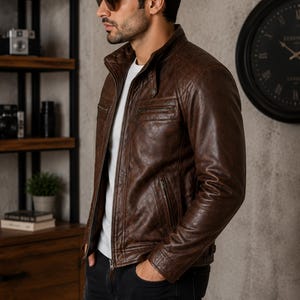 May include: A brown leather jacket featuring a stand-up collar, a front zipper closure, and quilted shoulder detailing. The jacket includes two zippered chest pockets and two lower zippered pockets, worn over a plain white t-shirt and black jeans.