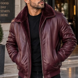 May include: A burgundy leather bomber jacket featuring a dark maroon faux fur collar, a front zipper closure, and ribbed cuffs and hem. The jacket is worn over a black sweater and black pants, with the wearer also sporting dark sunglasses.