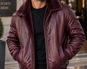 May include: A burgundy leather bomber jacket featuring a dark maroon faux fur collar, a front zipper closure, and ribbed cuffs and hem. The jacket is worn over a black sweater and black pants, with the wearer also sporting dark sunglasses.
