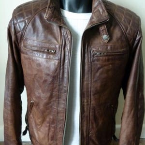 May include: A brown leather jacket featuring a quilted pattern on the shoulders, a stand-up collar with a snap button, and two horizontal zippered chest pockets. The jacket is worn over a white ribbed shirt and has a central full-length zipper.