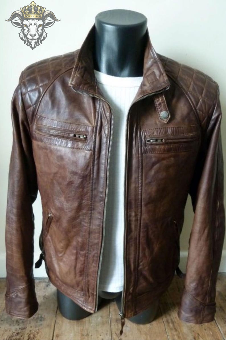 May include: A brown leather jacket featuring a quilted pattern on the shoulders, a stand-up collar with a snap button, and two horizontal zippered chest pockets. The jacket is worn over a white ribbed shirt and has a central full-length zipper.