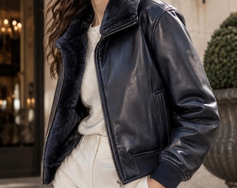 May include: A dark navy blue leather bomber jacket featuring a plush, dark-toned faux fur lining and collar, a central metal zipper closure, and ribbed knit cuffs. The jacket is worn over a cream-colored knit sweater and light-colored trousers.