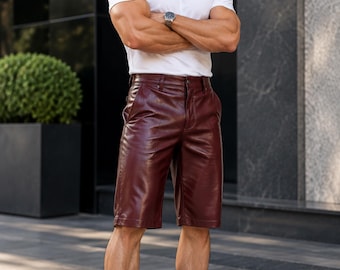 May include: A pair of burgundy leather shorts featuring a button and zipper fly closure, belt loops, and side pockets. The shorts have a smooth, semi-gloss finish and reach above the knee, paired with a plain white t-shirt and white sneakers.