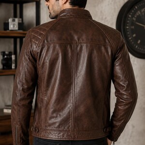 May include: A brown leather jacket featuring a stand-up collar, quilted shoulder panels, and long sleeves with zippered cuffs. The back of the jacket displays vertical seam detailing and adjustable side tabs at the hem, worn over black trousers.
