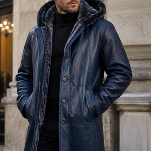 May include: A navy blue leather coat featuring a hood lined with dark gray faux fur, a button-front closure, and two side pockets. The coat is worn over a black turtleneck sweater and black trousers, providing a structured and layered appearance.