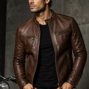 May include: A brown leather jacket featuring a mandarin collar with a snap button closure, a full-length front zipper, and two horizontal zippered chest pockets. The jacket has long sleeves with zippered cuffs and is worn over a black t-shirt.