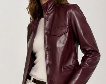 May include: A deep burgundy leather jacket featuring a high collar, long sleeves, and a front button closure. The jacket includes a single chest pocket with a flap and two lower pockets. It is worn over a white ribbed top and white pants with a dark belt.