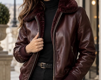 May include: A deep burgundy leather bomber jacket featuring a plush, matching faux fur collar, a central metal zipper closure, and ribbed knit cuffs and hem. The jacket is worn over a black turtleneck sweater and black trousers with a black belt.
