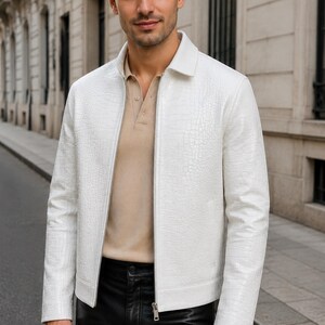 May include: A white jacket with a textured crocodile-skin pattern, a classic collar, and a full-length silver zip fastening. The jacket is worn over a beige polo shirt and black trousers, featuring long sleeves and a straight hemline.