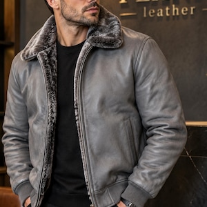 May include: A grey leather bomber jacket featuring a plush grey faux fur lining and a wide fold-over collar. The jacket includes a central metal zipper closure, ribbed cuffs at the sleeves and hem, and two side pockets. The exterior is smooth leather.