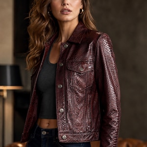 Puede incluir: Una chaqueta de color burdeos oscuro con un patrón texturizado de piel de serpiente, que presenta un cuello clásico, mangas largas y un cierre frontal con botones metálicos. La chaqueta incluye dos bolsillos en el pecho con solapas abotonadas.