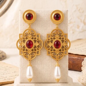 Könnte beinhalten: Ein Paar goldfarbene Ohrhänger mit einem runden Oberteil mit rotem Cabochon, einem quadratischen filigranen Mittelteil mit passendem rotem Cabochon und einer weißen, tropfenförmigen Kunstperle, die an der Unterseite herabhängt.