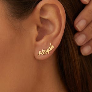 Puede incluir: Pendientes dorados con el nombre "Aliyah" escrito en cursiva. Los pendientes se llevan en una oreja, con una mano tocando suavemente la oreja. Joyería personalizada.