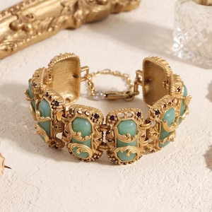May include: A gold-toned metal bracelet featuring rectangular links with inset light green stones. Each link is decorated with ornate scrollwork and small dark purple accents. The bracelet includes a chain extender with a lobster claw clasp.