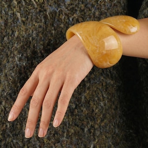 Puede incluir: Un brazalete amarillo-anaranjado con un patrón veteado. El brazalete se lleva en la muñeca, mostrando su diseño curvo. La joya tiene un acabado brillante y está hecha de un material sólido. El fondo es una tela texturizada de color verde oscuro y marrón.