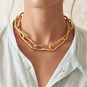 May include: A gold-toned chain necklace featuring large, elongated, oval-shaped links with a textured, uneven surface. The necklace rests around the neck, showcasing a chunky, bold design with a metallic finish and a consistent link pattern throughout.
