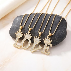 May include: Gold-toned necklaces spelling "LOVE", each featuring a crown-topped initial pendant encrusted with clear crystals. The pendants are suspended from delicate gold chains, displayed on a dark gray stone.