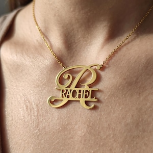 May include: A gold-colored necklace with a pendant featuring the name "RACHEL" in a stylized font. The pendant includes a large, decorative initial "R" above the name, all suspended from a delicate chain.