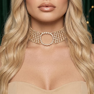 Op de afbeelding: Een goudkleurige choker-halsketting met een brede, gaasachtige band bezet met talloze heldere, sprankelende strass-steentjes. Het midden van de choker heeft een opvallende, ronde open uitsparing omlijst door een ring van bijpassende strass-steentjes.