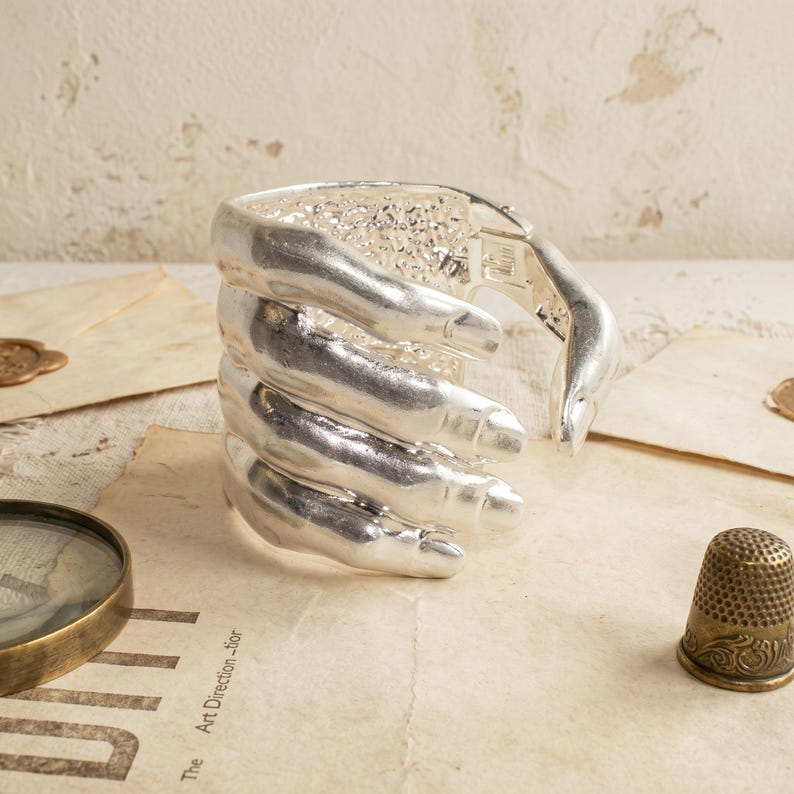 May include: A silver-toned cuff bracelet shaped like a hand wrapping around the wrist. The design features realistic finger detailing with a smooth, polished metallic finish on the exterior and a textured, rough surface on the inner band.