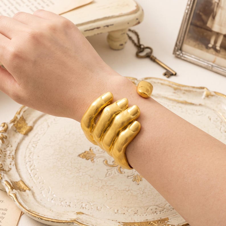 May include: A gold-toned cuff bracelet shaped like a hand wrapping around the wrist. The design features smooth, metallic fingers that curve to form the band, with a polished finish and a small, rounded terminal end on the opposite side.