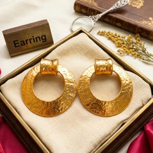 May include: A pair of gold-toned earrings featuring a large, circular hoop design with a textured, hammered surface. Each earring has a smooth, cylindrical top connector piece. The word Earring is printed on a small wooden block nearby.
