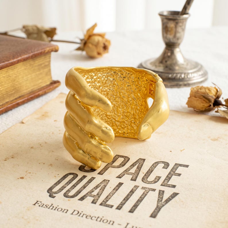 May include: A gold-toned cuff bracelet featuring a sculptural design of fingers wrapping around the wrist. The band has a textured, hammered finish on the interior surface. The bracelet rests on a page with the printed text SPACE QUALITY.