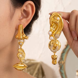 May include: A pair of gold-toned earrings featuring anatomical motifs. One earring depicts a nose, a sunburst, a coiled shape, and lips, all accented with clear crystals. The other earring is shaped like an ear with pearl and crystal embellishments.