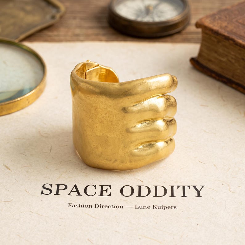 May include: A gold-toned cuff bracelet shaped like a stylized hand with fingers wrapped around the wrist. The surface has a textured, matte finish. Below the bracelet, the text reads SPACE ODDITY, Fashion Direction &mdash; Lune Kuipers.