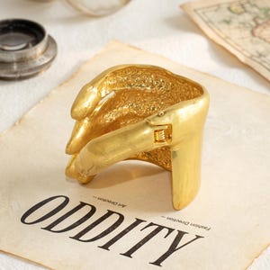 May include: A gold-toned metal cuff bracelet shaped like a hand with a textured palm and smooth fingers. The bracelet features a hinged mechanism on the side, allowing it to open and close securely around the wrist with a polished finish.