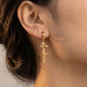 May include: Gold-colored hoop earring with the name "Andrea" in a cursive script. The earring is attached to a person's ear. The earring is a simple, elegant piece of jewelry.
