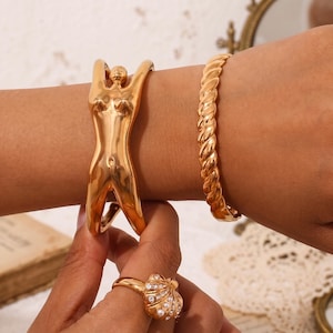 Op de afbeelding: Twee goudkleurige armbanden en een goudkleurige ring. De ene armband heeft een gestileerd ontwerp van een menselijke figuur, terwijl de tweede armband een gedraaide touwstructuur heeft. De ring is schelpvormig en bezet met kleine doorzichtige steentjes.