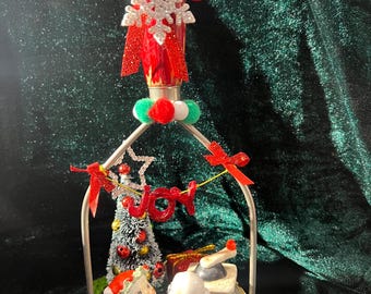 Puede incluir: Un machacador de cocina decorativo con mango rojo rematado por un copo de nieve plateado brillante y un lazo rojo. El marco de metal contiene un árbol de Navidad en miniatura, dos figuras pequeñas en rojo y una pancarta con el texto JOY.