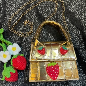 May include: A gold-colored handbag with a chain strap and a strawberry design. The bag has a crocodile-embossed texture and a top handle shaped like a strawberry.