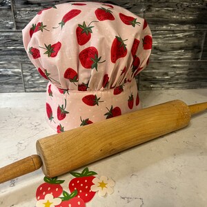 Puede incluir: Un gorro de chef rosa con estampado de fresas y lunares blancos. Un rodillo de madera y decoraciones con forma de fresa est&aacute;n sobre una encimera blanca.