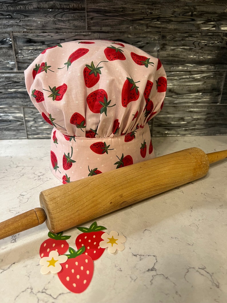 Puede incluir: Un gorro de chef rosa con estampado de fresas y lunares blancos. Un rodillo de madera y decoraciones con forma de fresa est&aacute;n sobre una encimera blanca.