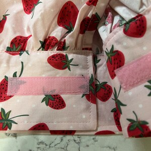 Puede incluir: Un gorro de chef de tela rosa con un patr&oacute;n de lunares blancos y estampado de fresas rojas. El gorro tiene correas ajustables rosas con cierres de velcro.