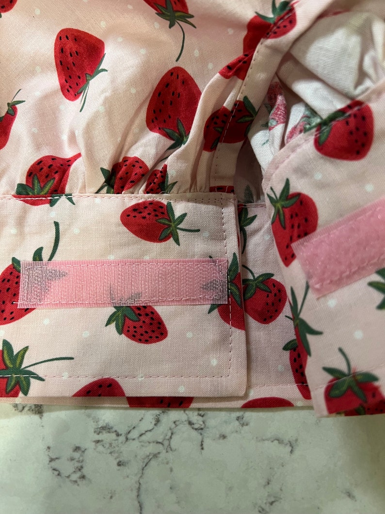 Puede incluir: Un gorro de chef de tela rosa con un patr&oacute;n de lunares blancos y estampado de fresas rojas. El gorro tiene correas ajustables rosas con cierres de velcro.