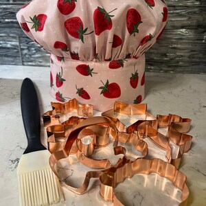 Puede incluir: Un gorro de chef rosa con estampado de fresas y un juego de cortadores de galletas de cobre. Tambi&eacute;n se incluye un pincel de reposter&iacute;a de silicona.