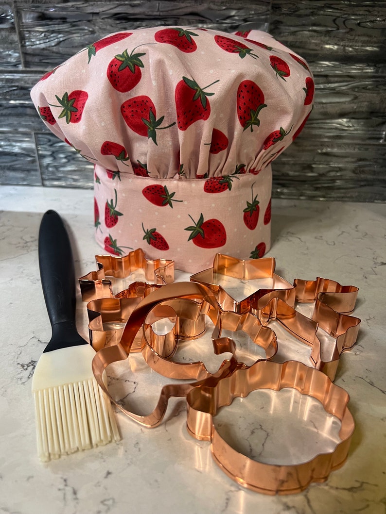 Puede incluir: Un gorro de chef rosa con estampado de fresas y un juego de cortadores de galletas de cobre. Tambi&eacute;n se incluye un pincel de reposter&iacute;a de silicona.