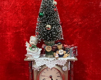 Puede incluir: Un reloj de repisa decorativo con esfera dorada y números romanos, adornado con detalles de pedrería. Sobre el reloj hay un árbol de Navidad en miniatura con adornos, un ángel de cerámica, un pequeño oso de peluche y un regalo.