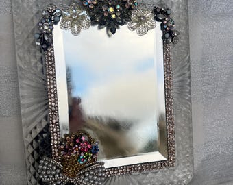 Puede incluir: Un espejo rectangular de cristal con un borde de patrón de diamantes. El marco está adornado con pedrería iridiscente, detalles florales metálicos y un grupo de gemas coloridas con forma de cesta con un lazo de perlas blancas.