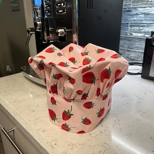 Puede incluir: Un gorro de chef rosa y blanco con estampado de fresas. El gorro est&aacute; sobre una encimera blanca.