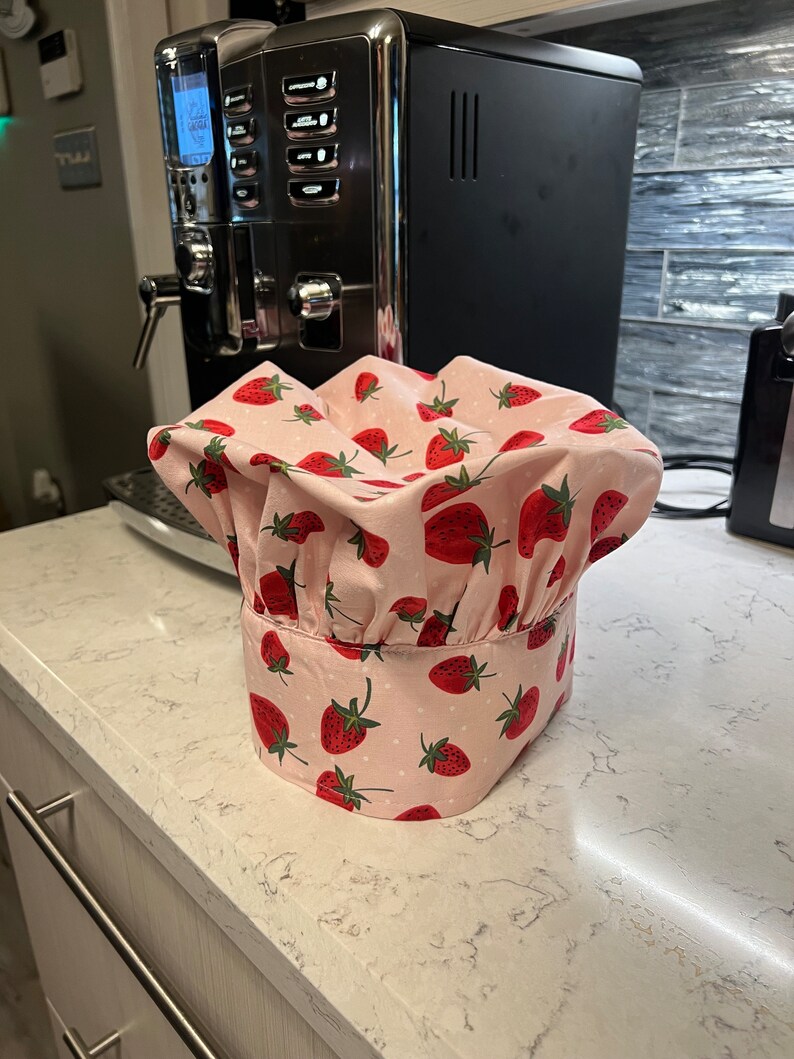 Puede incluir: Un gorro de chef rosa y blanco con estampado de fresas. El gorro est&aacute; sobre una encimera blanca.