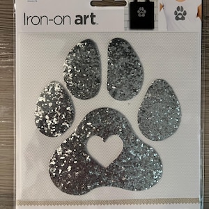 May include: Silver glitter iron-on paw print art with a heart cutout. The package reads "Iron-on art" and "quick and easy". The image shows a tote bag and a t-shirt with paw print designs.